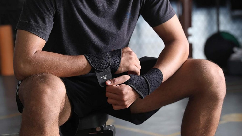 A man pulls the strap of a wrist brace to secure it while sitting in a gym.