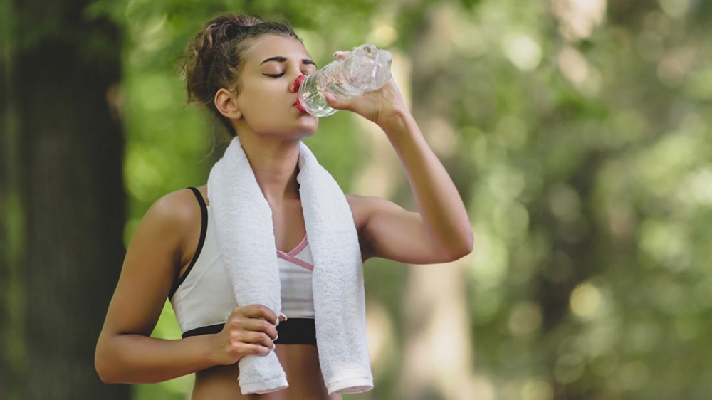 A young woman drinking water and cooling off with a white towel around her neck after her running at the park.